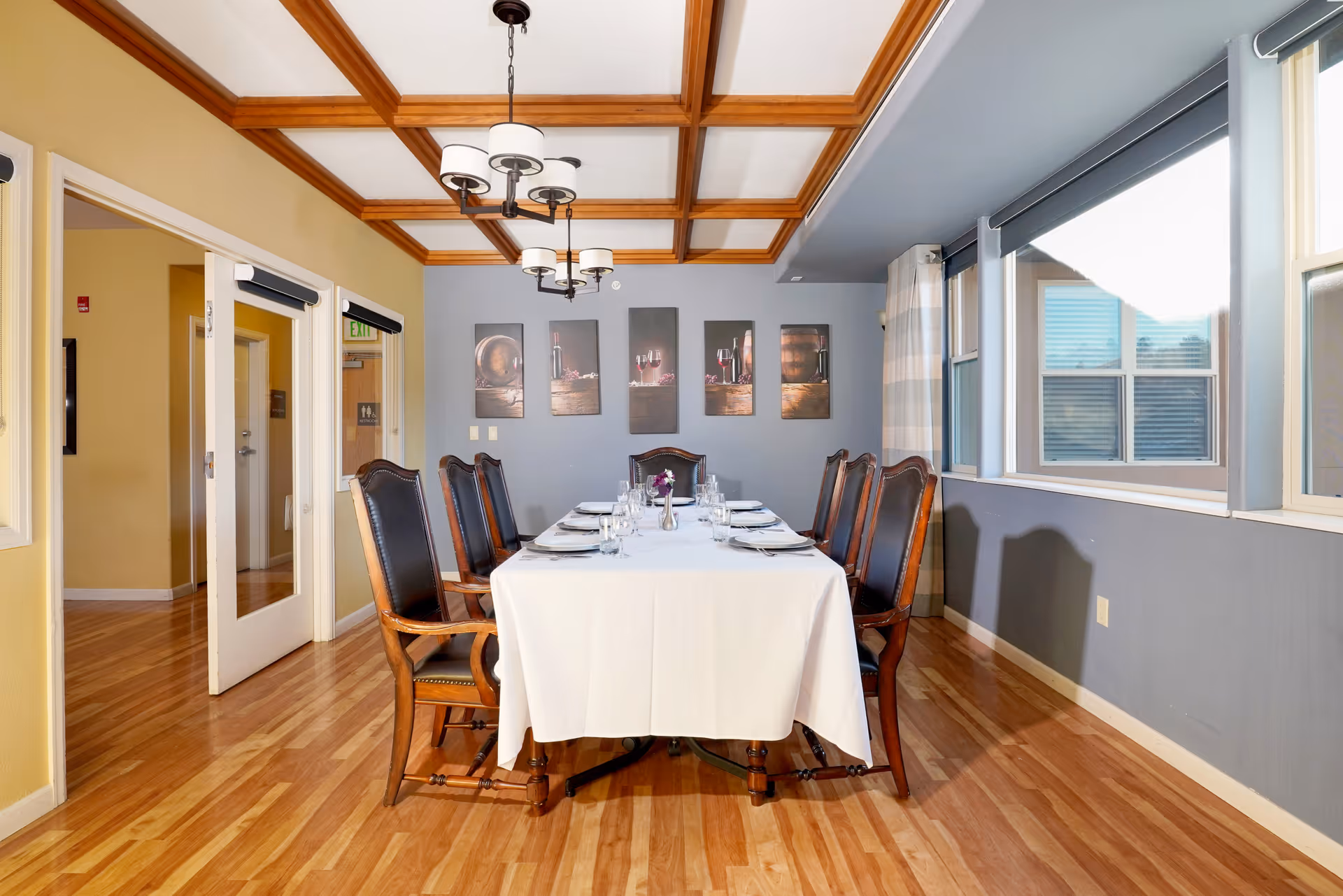 A formal dining room with a long rectangular table covered with a white tablecloth, set with plates, glasses, and silverware. The table is surrounded by eight wooden chairs with dark leather upholstery. The room has wooden flooring, a coffered ceiling with wooden beams, and a chandelier with multiple lights. On the far wall, there are five framed pictures, and large windows with blinds and curtains are on the right side.