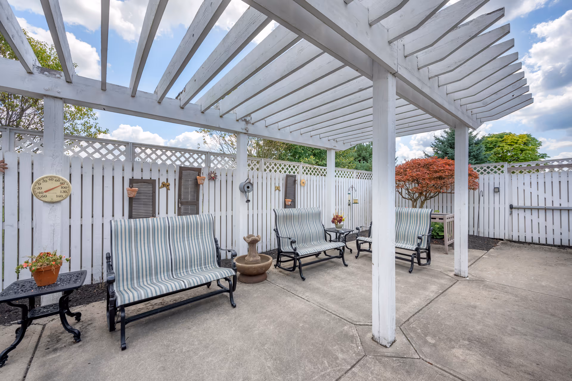 Outdoor patio area with a white wooden pergola overhead, three striped cushioned benches, a small black table with a potted plant, a decorative water fountain, and a white picket fence with lattice top surrounding the space. There are some small decorative items on the fence and a tree with red leaves in the background under a partly cloudy sky.