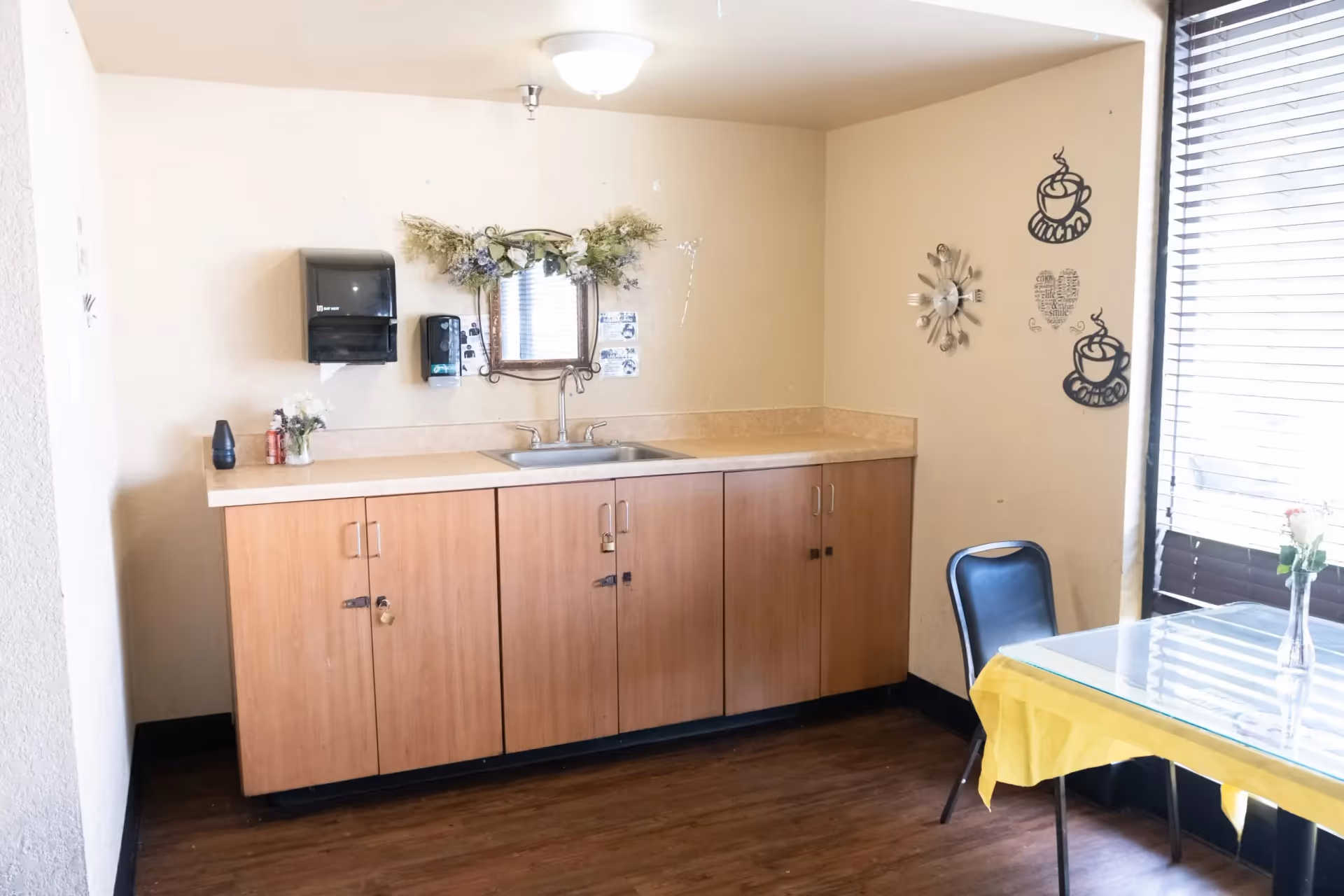 A small kitchenette area with wooden cabinets, a countertop with a sink, a wall-mounted paper towel dispenser, and a soap dispenser. Above the sink is a mirror decorated with a floral arrangement. To the right, there is a table with a yellow tablecloth and a single black chair. The wall has decorative coffee cup and heart-shaped wall art, and a window with blinds lets in natural light.