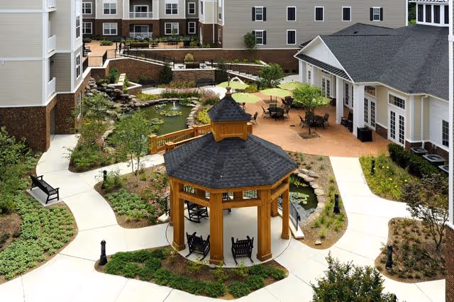 A landscaped courtyard with a central wooden gazebo, pond, patio seating, and surrounding multi-story senior living buildings.
