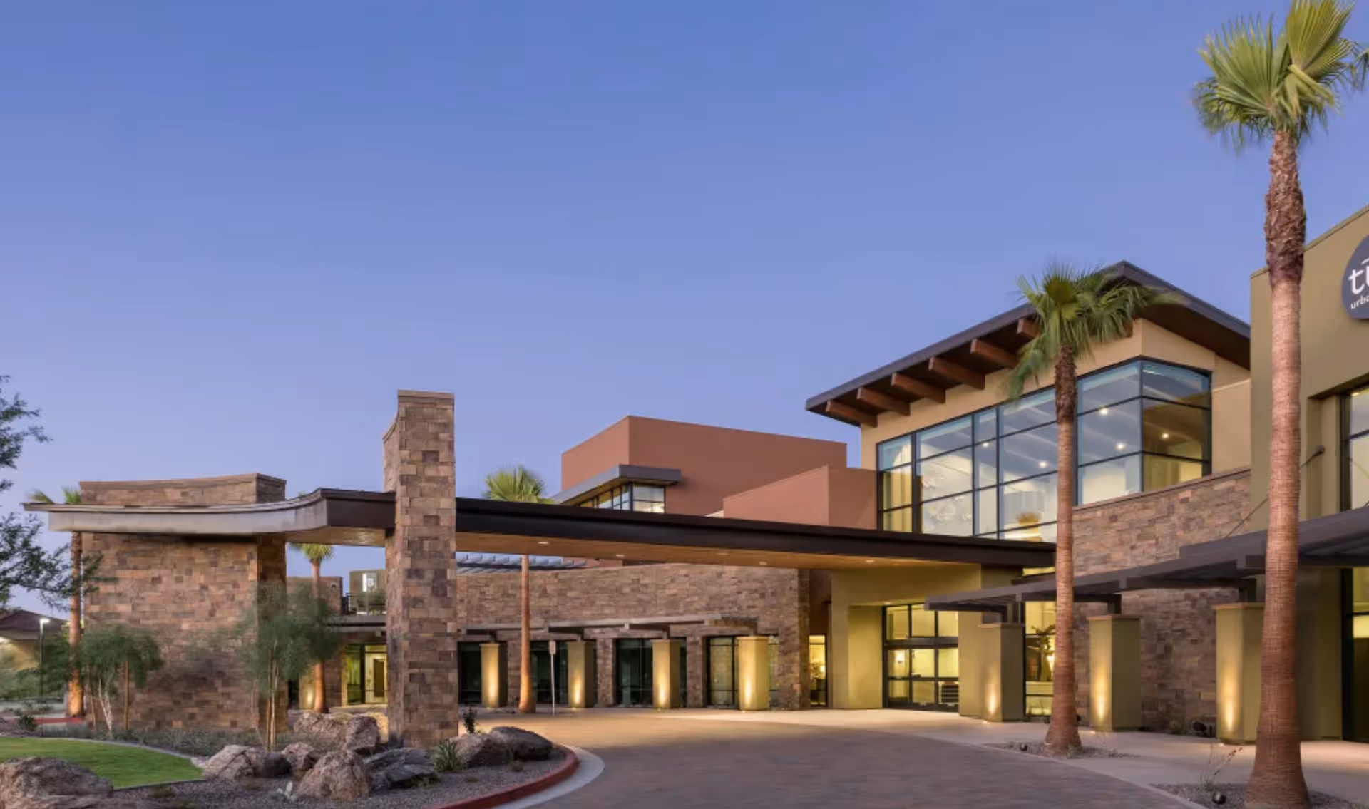 Exterior view of a modern building at dusk with stone and stucco walls, large windows, palm trees, and a covered driveway entrance.