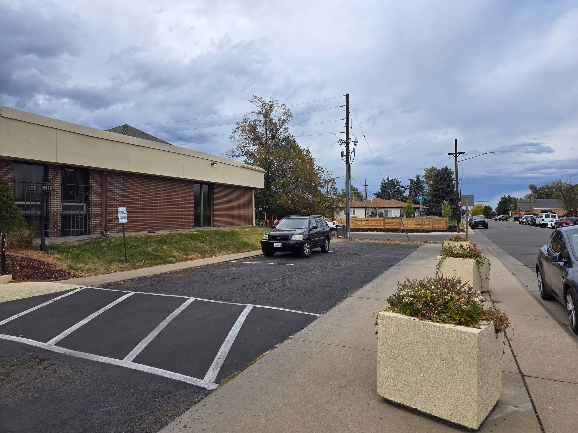 Exterior view of a single-story brick building with a beige roof trim, adjacent to a parking lot with a few parked cars. The sky is cloudy, and there are two large rectangular planters with plants on the sidewalk in the foreground. Residential houses and trees are visible in the background.