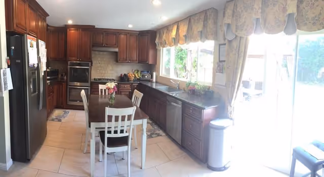 A bright kitchen with dark wooden cabinets, stainless steel appliances including a refrigerator, oven, and dishwasher. There is a small dining table with four white chairs in the center, a vase with flowers on the table, and large windows with floral valances letting in natural light. A sliding glass door leads to an outdoor area.