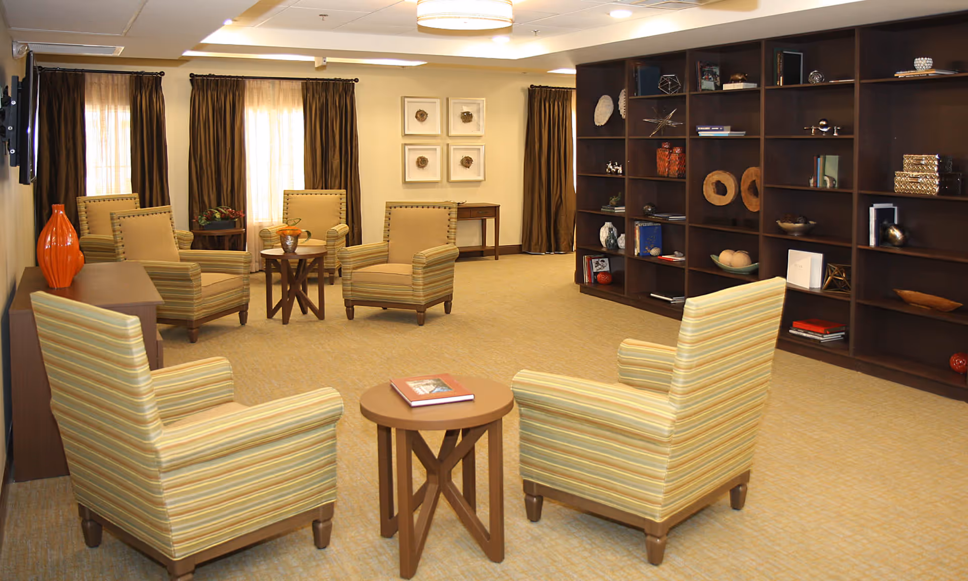A cozy living room area with six striped armchairs arranged around three small wooden tables. The room features beige carpeting, dark brown curtains on windows, a large dark wooden bookshelf filled with decorative items and books, and framed artwork on the wall.