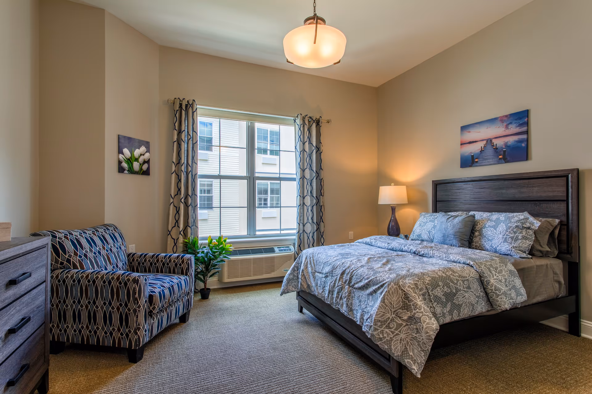 A cozy bedroom with a large window covered by patterned curtains, a bed with floral bedding and multiple pillows, a bedside table with a lamp, a patterned armchair, a dresser, and two framed pictures on the walls.