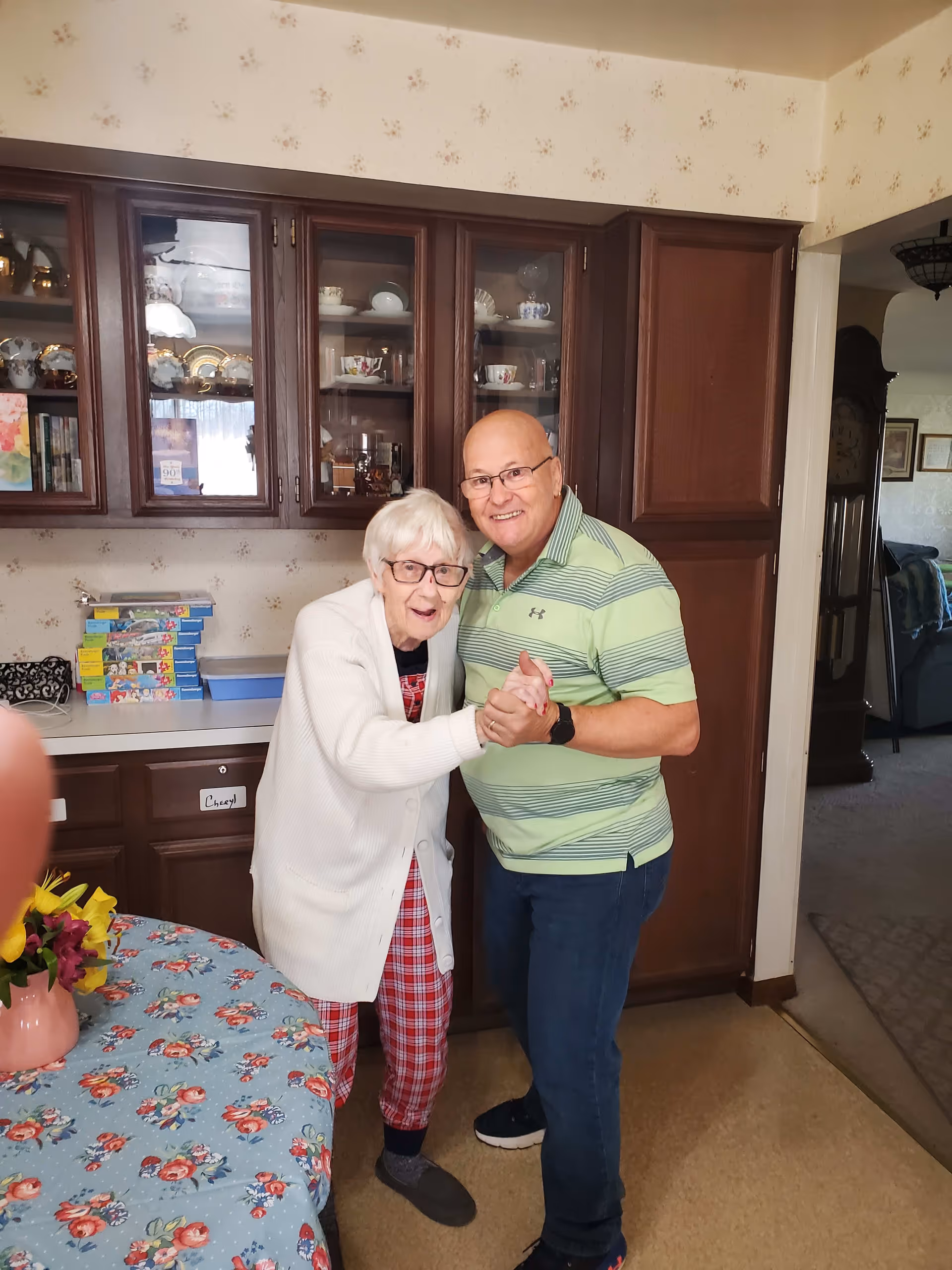 An elderly woman wearing glasses, a white cardigan, and red plaid pants is holding hands and smiling with a middle-aged man in a green striped polo shirt and glasses. They are standing in a kitchen with dark wooden cabinets and a table covered with a floral tablecloth and a vase of flowers.