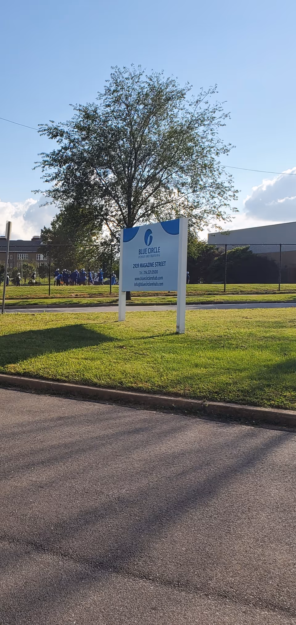 Outdoor view of a grassy area with a large tree and a sign for Blue Circle Rehab and Nursing. The sign displays the facility's name, address, phone number, and website. In the background, there is a fence, some buildings, and a group of people gathered near the fence.