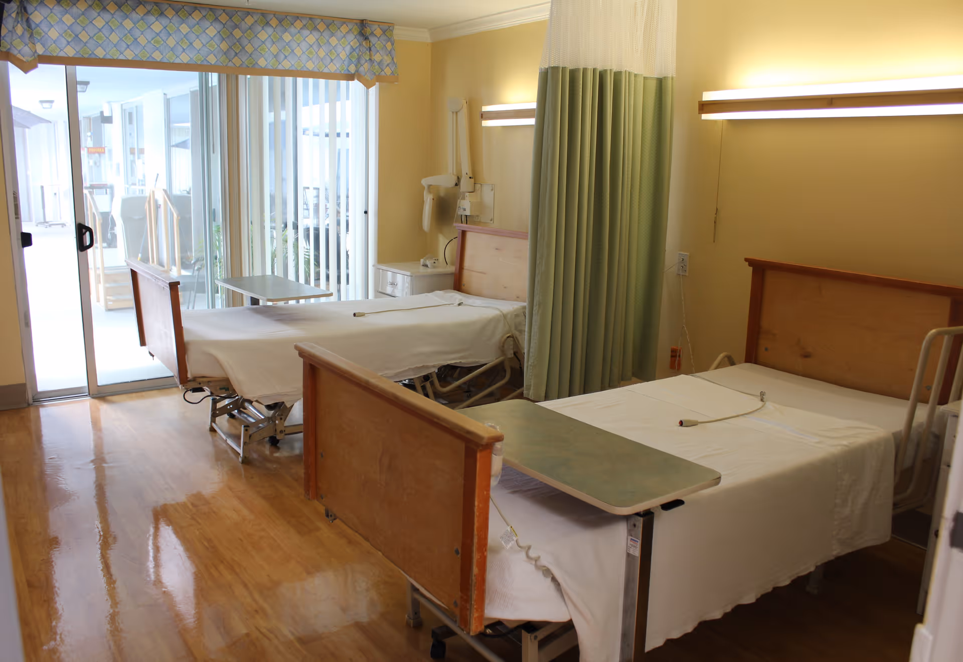 A room with two hospital-style beds separated by a green and white privacy curtain. Each bed has a white sheet and an overbed table. The room has wooden floors, yellow walls, and a sliding glass door leading to an outdoor area. There are wall-mounted lights above each bed and medical equipment attached to the wall near one bed.