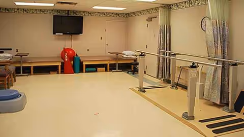 A rehabilitation therapy room with parallel bars, mats, curtains, benches, and a wall-mounted TV.