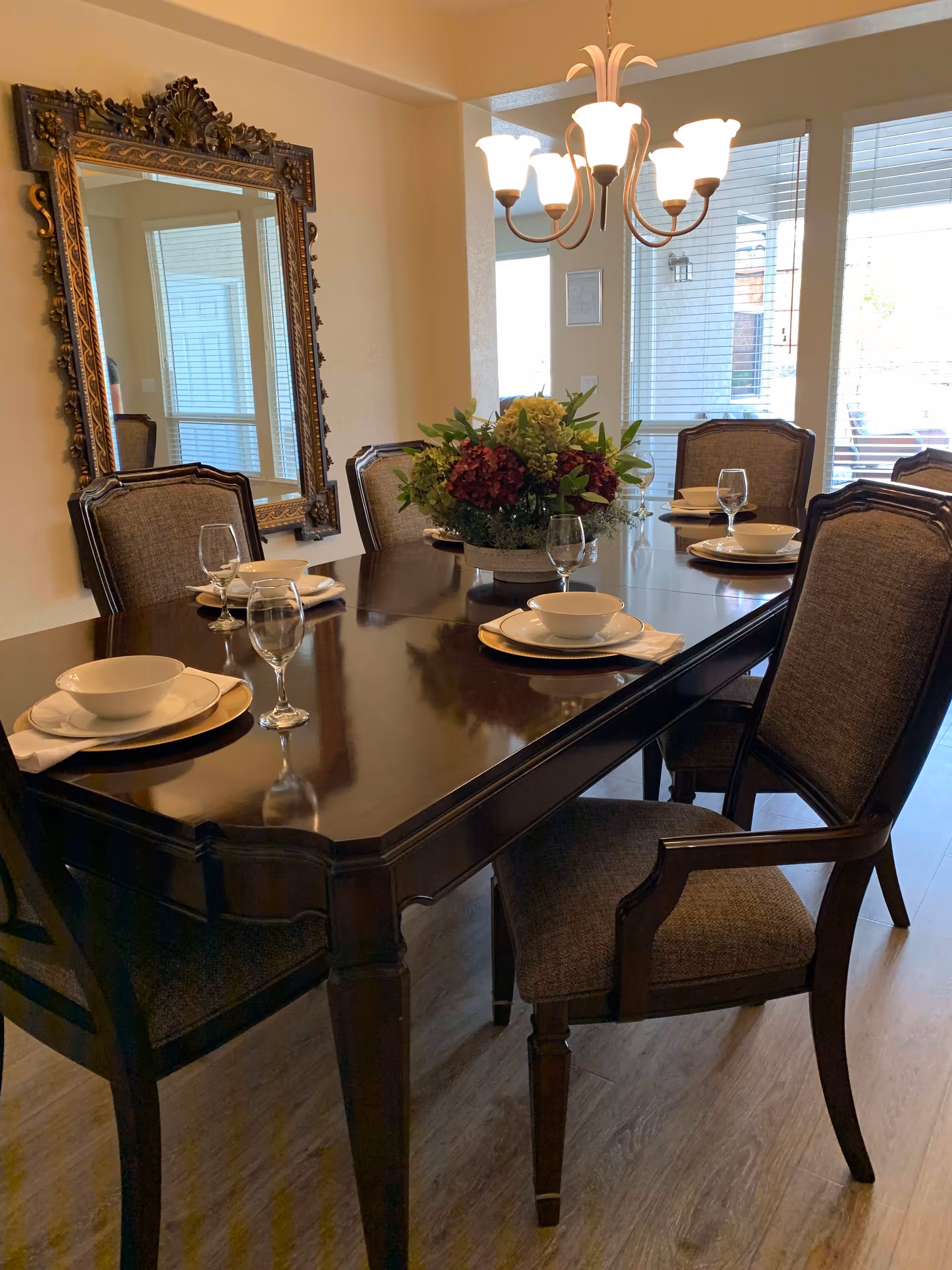 A dining room with a dark wooden table set for six people, each place setting includes a white bowl, plate, napkin, and a wine glass. There is a floral centerpiece on the table. The room features a large ornate mirror on the wall and a chandelier with multiple lights hanging above the table. Large windows with blinds allow natural light into the room.