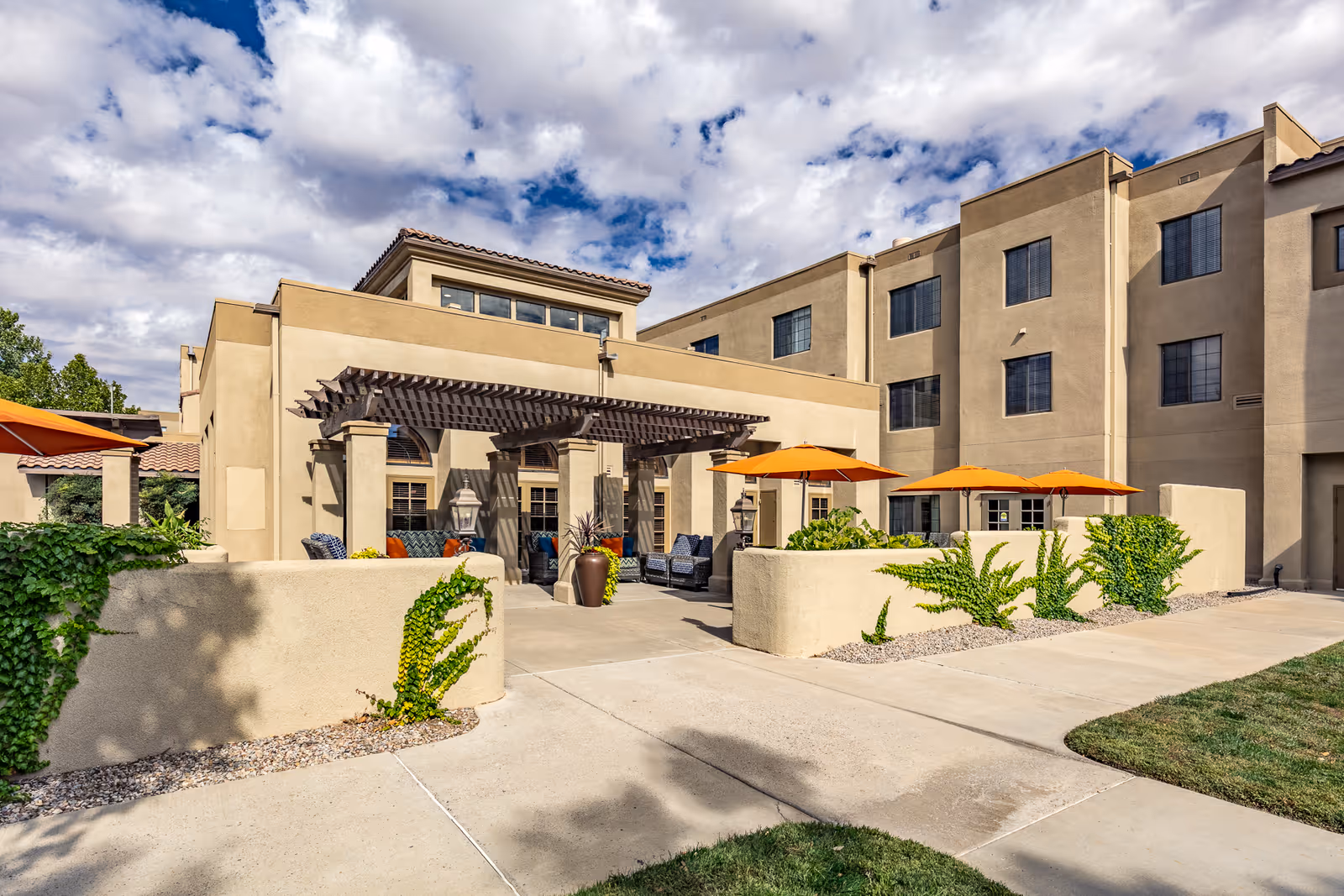Outdoor patio area of a senior living facility with beige stucco walls, orange umbrellas shading seating areas, green plants, and a pergola over a seating space. The building is multi-story with several windows and a partly cloudy sky above.