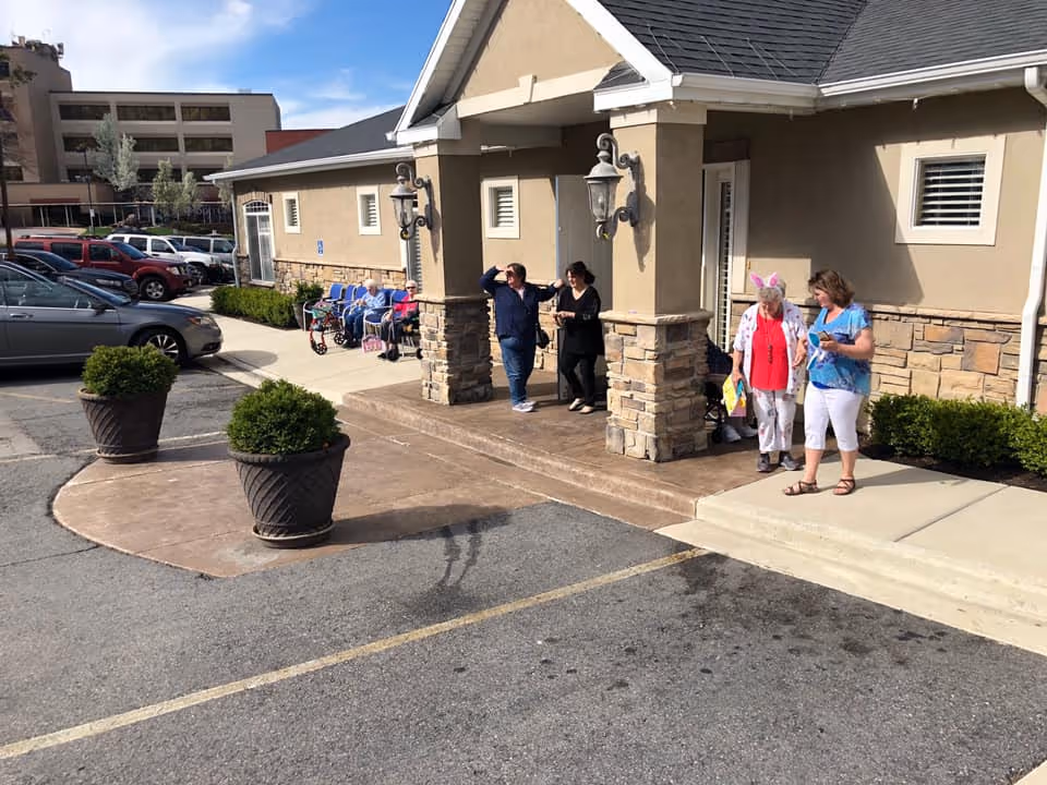 Exterior view of the entrance to Welcome Home Assisted Living facility with a covered porch supported by stone pillars. Several people, including elderly individuals and caregivers, are standing and sitting near the entrance. There are large potted plants on the concrete area in front of the entrance and parked cars visible in the background.