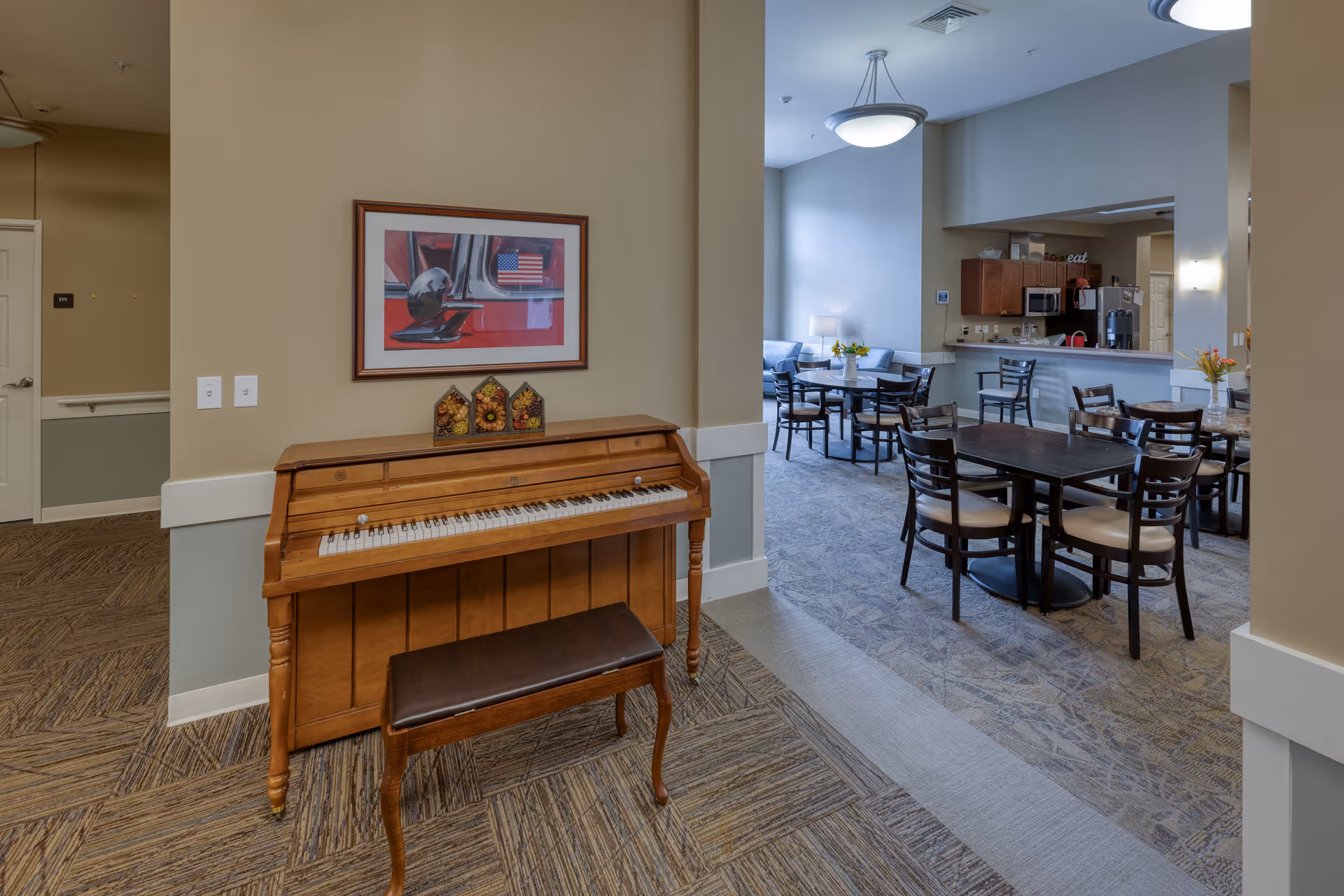 An upright wooden piano and bench sit near a communal dining area with tables, chairs and a serving counter.
