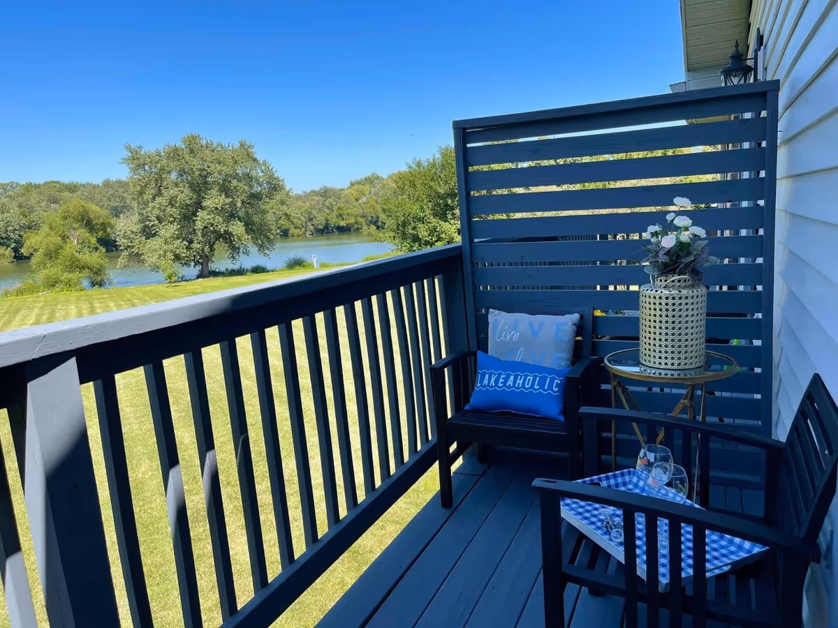 A small balcony with dark wooden railings and floor, featuring two black chairs with blue cushions, a small round table with a decorative vase of flowers, overlooking a grassy area and a river with trees in the background under a clear blue sky.