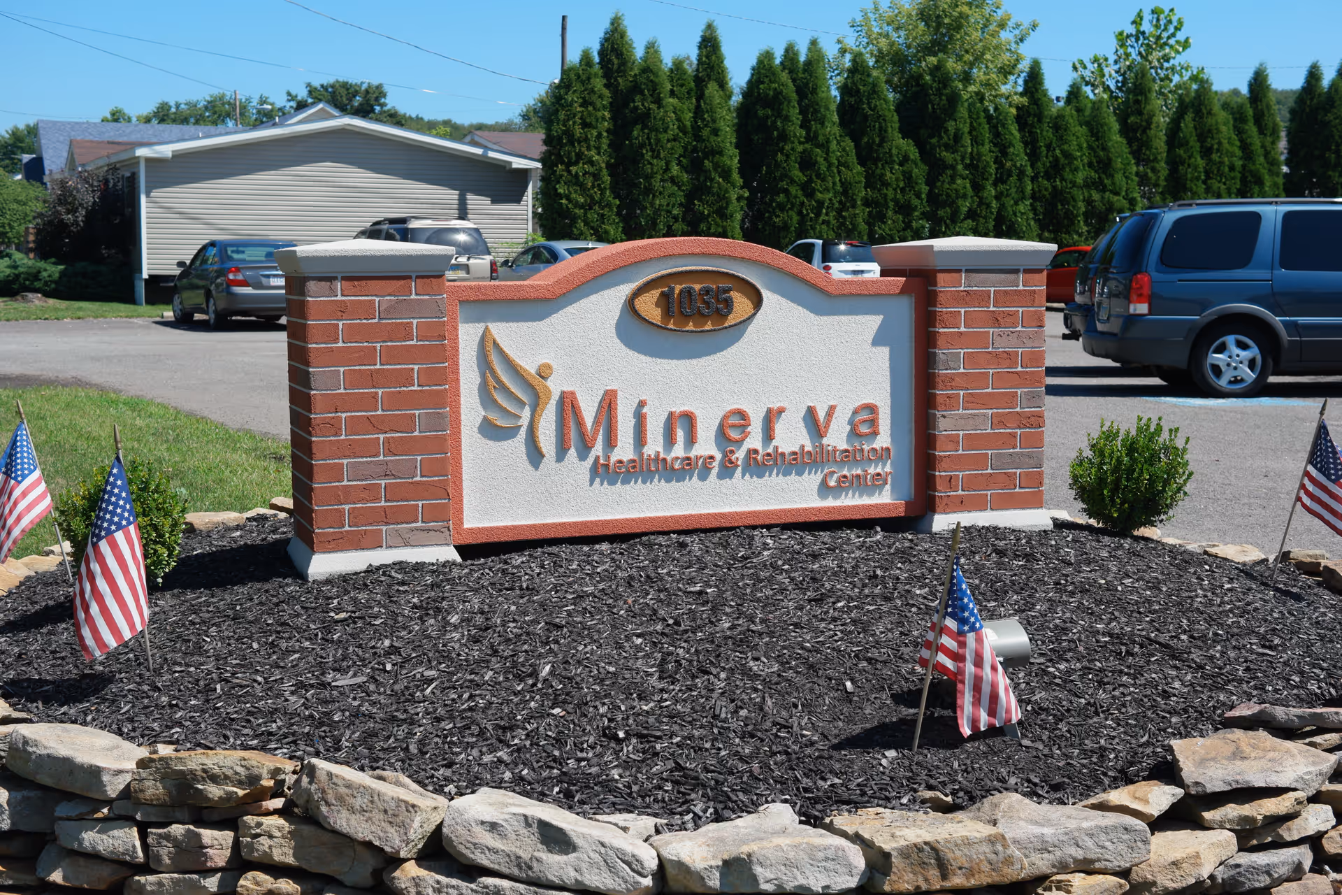 Brick and stone entrance sign for Minerva Healthcare & Rehabilitation Center with small American flags and parked cars in the background.