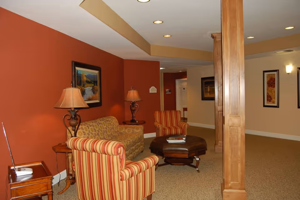 Cozy seating area with striped armchairs, a patterned sofa, ottoman and lamps against a red accent wall in a common room.