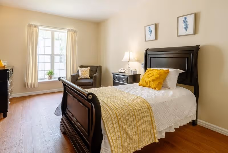 A bright bedroom with a single dark wooden bed featuring white bedding and a yellow throw blanket and pillow. There is a dark wooden nightstand with a lamp and a phone, a dark armchair with a decorative pillow, and a large window with cream curtains letting in natural light. Two framed leaf prints hang on the beige wall above the bed.