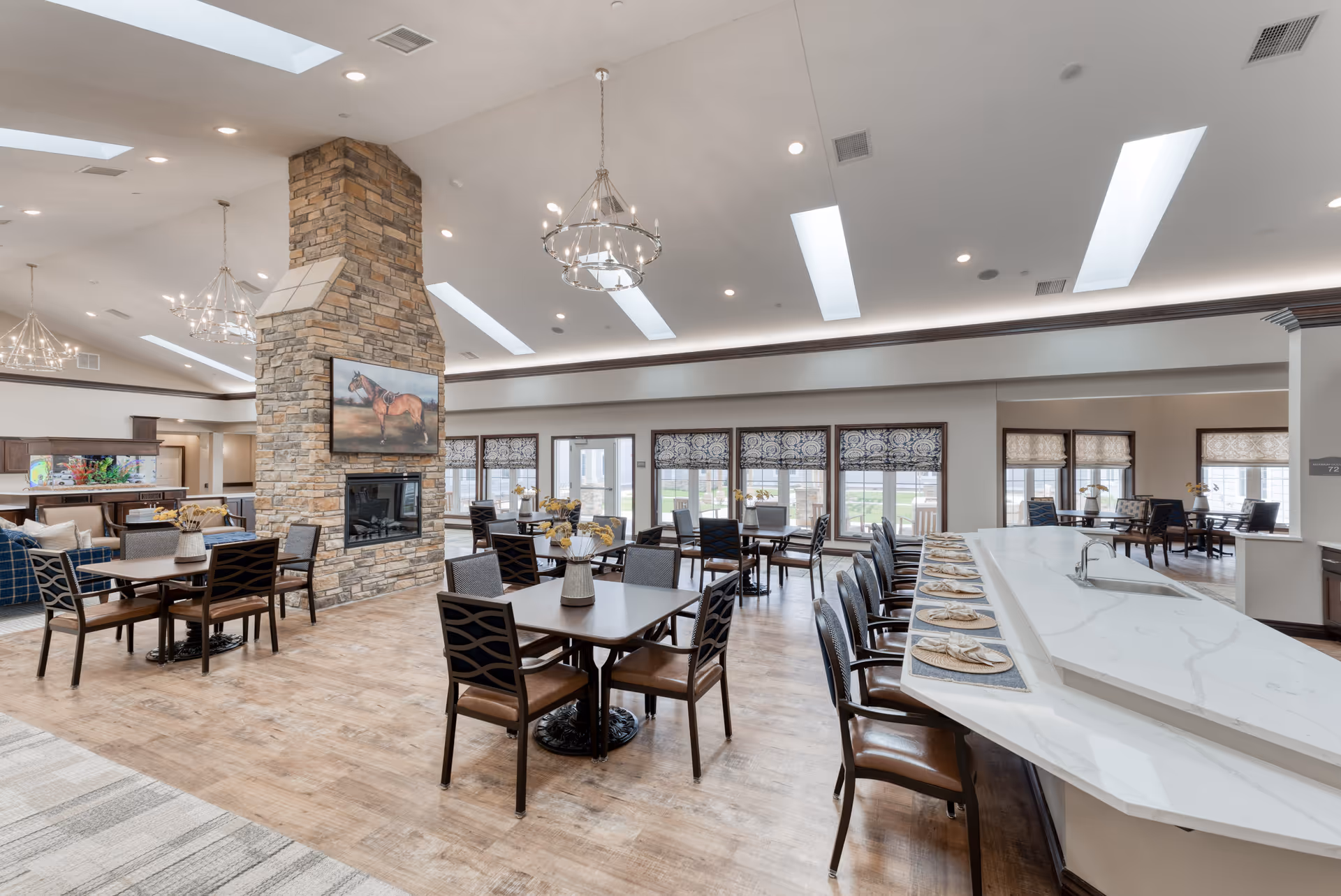 A spacious senior living dining area with multiple tables and chairs, a large stone fireplace with a painting of a horse above it, and a long white marble counter set with placemats and napkins. The room features skylights, chandeliers, large windows with patterned shades, and wooden flooring.