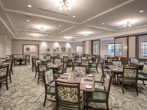 A spacious dining room with multiple wooden tables and chairs arranged neatly. The chairs have floral patterned upholstery and green cushions. The room features a patterned carpet, recessed ceiling lights, and decorative wall art. Large windows with wooden lattice frames allow natural light to enter.