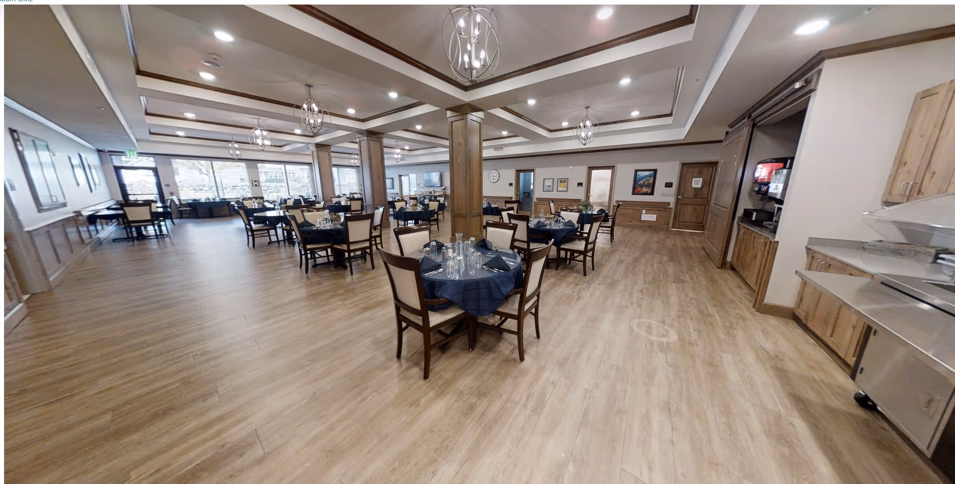 Spacious dining room in Whisper Cove Assisted Living and Memory Care with multiple round tables covered with dark blue tablecloths, each set with glassware and napkins. The room features wooden flooring, wooden columns, and modern ceiling light fixtures. Large windows on one side allow natural light to fill the space.