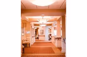 A well-lit hallway in a retirement community with beige walls, white trim, and a patterned carpet. The hallway features ceiling lights, framed artwork on the walls, and handrails along both sides. There are doorways and seating areas visible along the corridor.