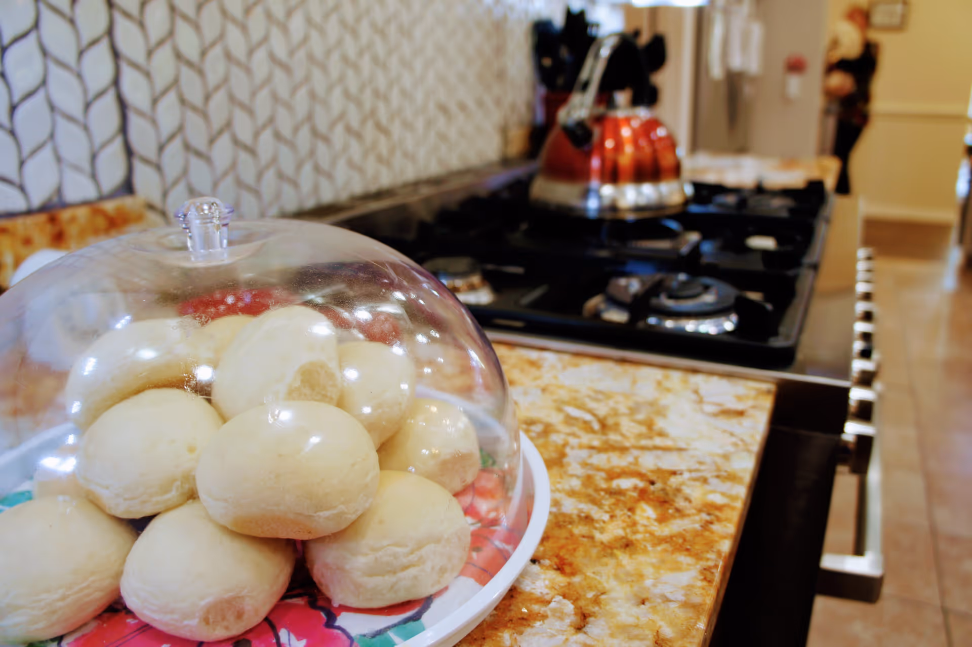 Close-up view of a kitchen countertop with a plate of bread rolls covered by a clear plastic dome in the foreground. In the background, there is a stove with a red kettle on it and a refrigerator further back in the room.