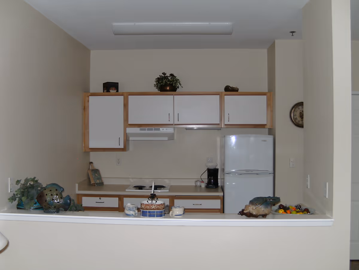 Small kitchen area with white upper and lower cabinets, a white refrigerator, a coffee maker on the counter, and a small stovetop. Decorative items including plants and ceramic pieces are placed on the counter and above the cabinets. The walls are beige and there is a clock on the right wall.