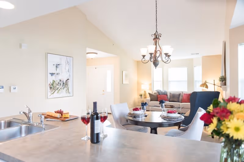 Bright and airy open-concept living space featuring a kitchen counter with a bottle of wine and two glasses, a round dining table set with plates and napkins, and a cozy living room with a sofa, armchair, and decorative pillows. A bouquet of flowers is visible in the foreground on the right side.