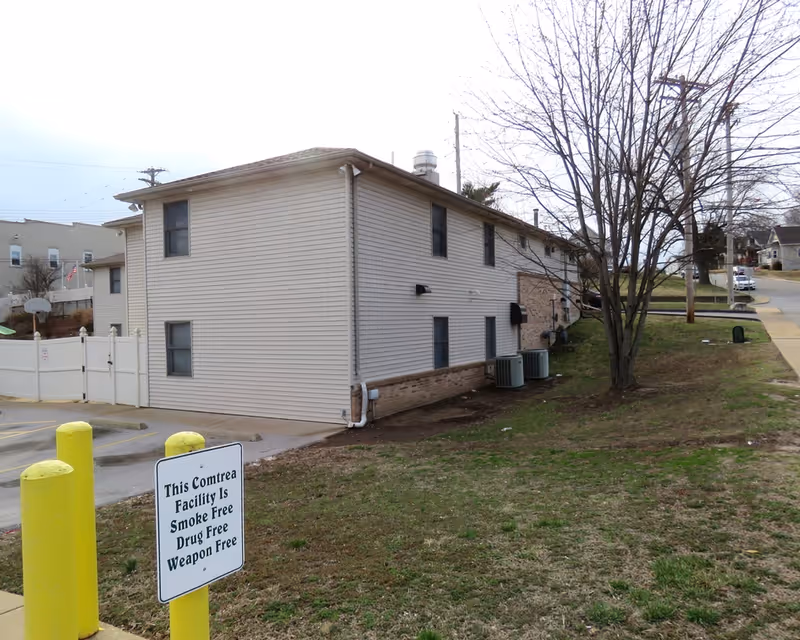 Exterior view of a two-story beige siding building with a small lawn, a leafless tree, HVAC units, and a sign noting the facility is smoke-, drug- and weapon-free.