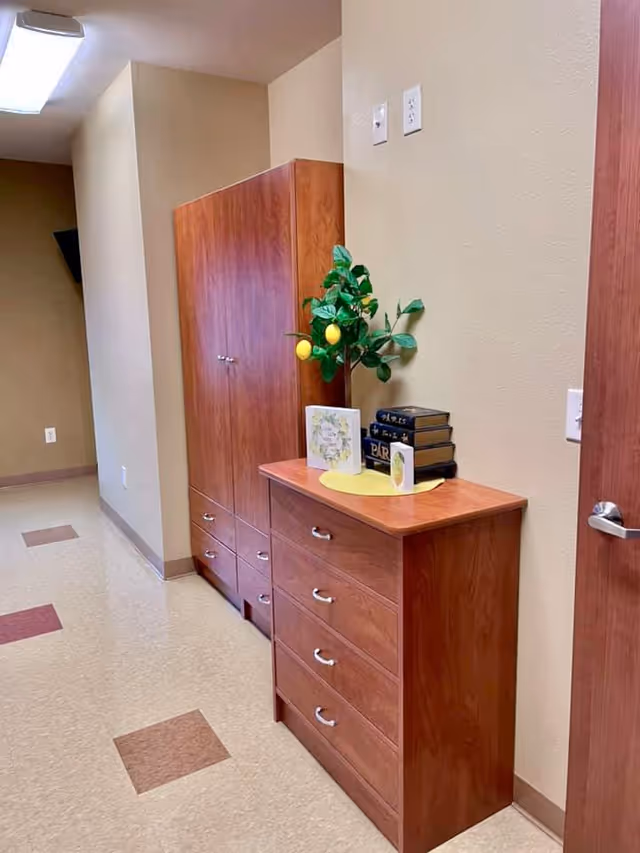A hallway corner in a senior living facility with wooden cabinets and drawers. On top of the drawers, there is a small decorative plant with yellow lemons, two greeting cards, and a stack of books. The walls are beige, and the floor has a pattern of beige and brown tiles.