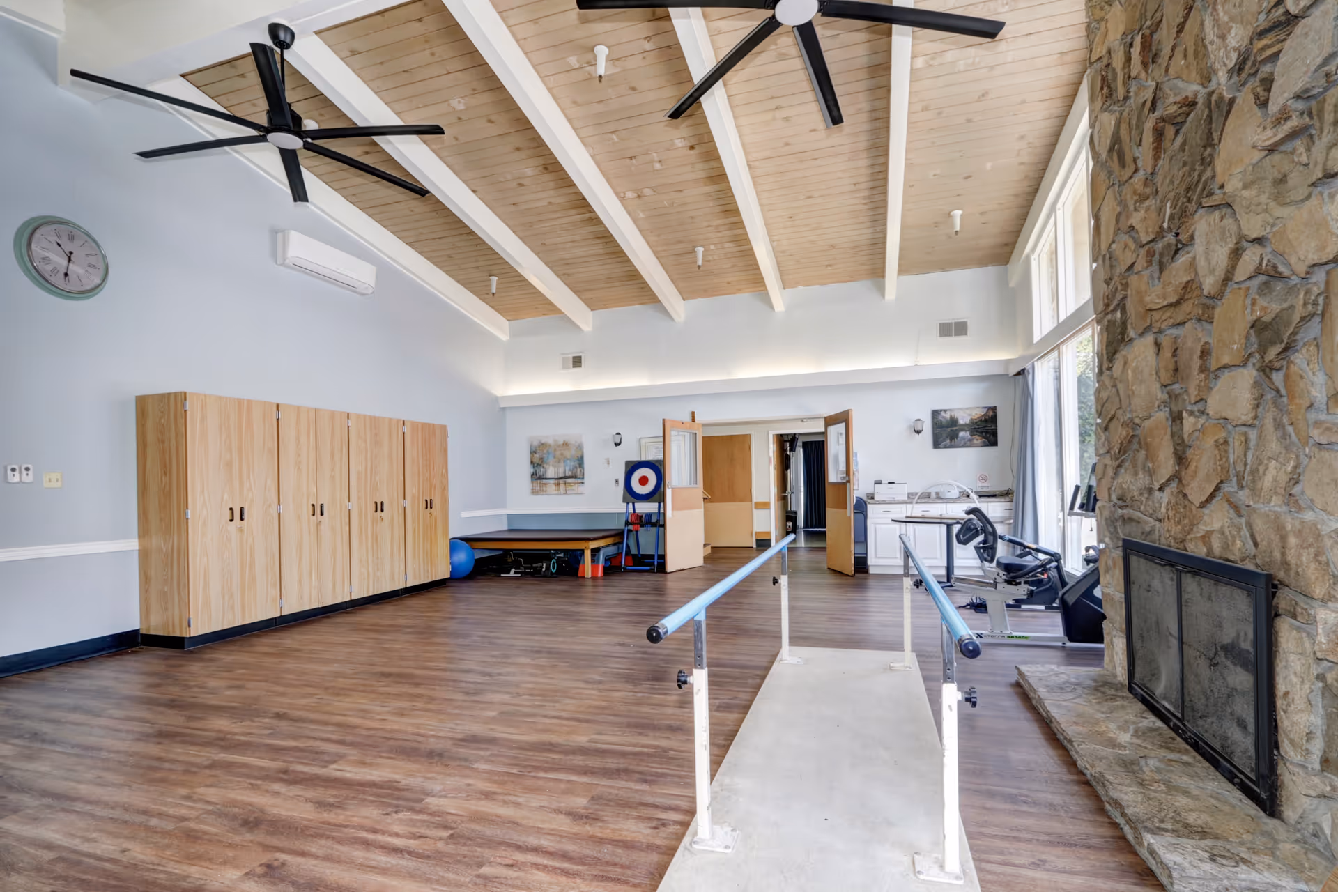 Large interior therapy/exercise room with parallel walking bars, workout equipment, wooden storage cabinets, and a stone fireplace under a beamed ceiling.