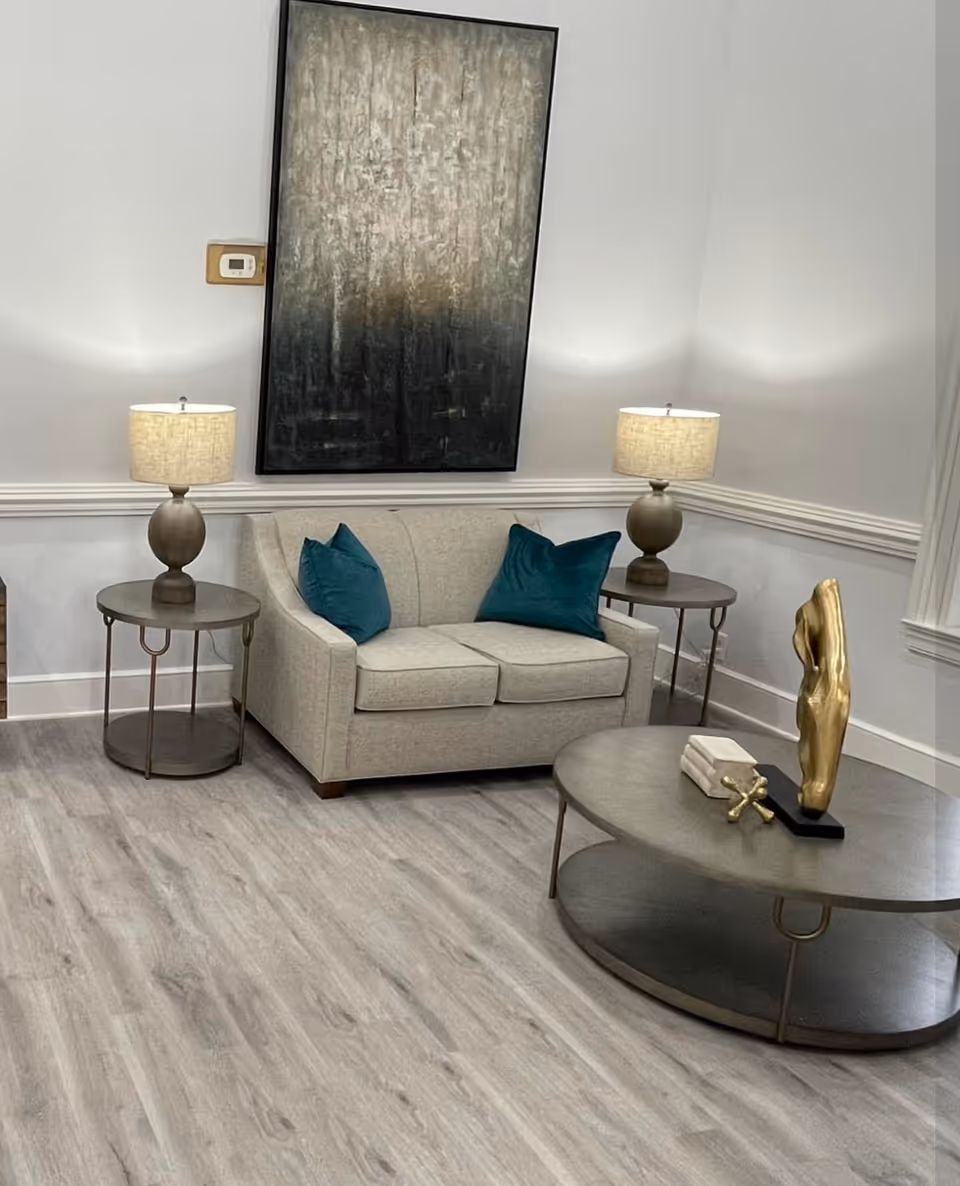 A small living room with a loveseat flanked by two lamps and side tables, a round coffee table with decorative sculpture, and abstract wall art above.