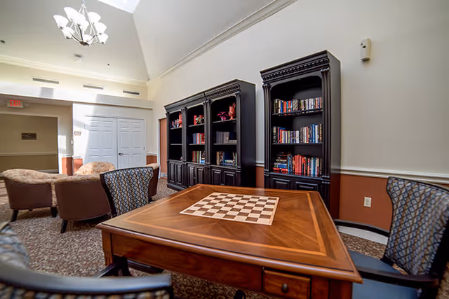 A cozy interior common area with a wooden table featuring a built-in chessboard, surrounded by patterned chairs. In the background, there are two tall black bookshelves filled with books and decorative items. Comfortable armchairs are arranged near a wall with white double doors, and a chandelier hangs from the ceiling.