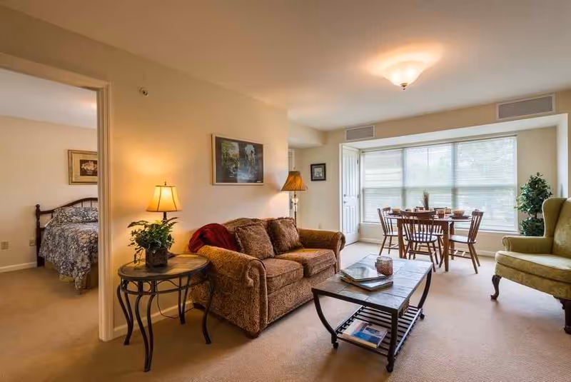 A cozy living room with a brown patterned sofa, a side table with a lamp and plant, a coffee table with books and a candle, and a green armchair. In the background, there is a dining area with a wooden table and chairs near a large window with blinds. A bedroom with a bed and floral bedspread is visible through an open doorway.