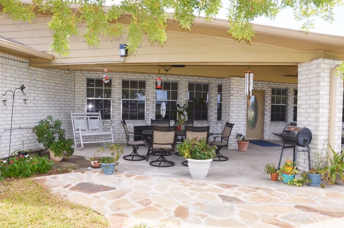 Covered outdoor patio area with a stone floor leading to a concrete seating area. The patio features a white rocking bench, a round table with six chairs, several potted plants, a hanging wind chime, and a charcoal grill. The building has light-colored brick walls and multiple windows with white frames.