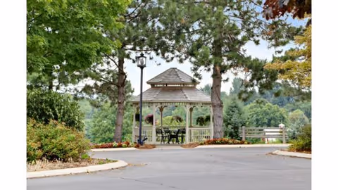 A wooden gazebo with a shingled roof situated between two tall pine trees, surrounded by greenery and bushes. There is a paved pathway leading up to the gazebo, which contains a table and chairs inside. The background shows a lush, green landscape with trees and hills.