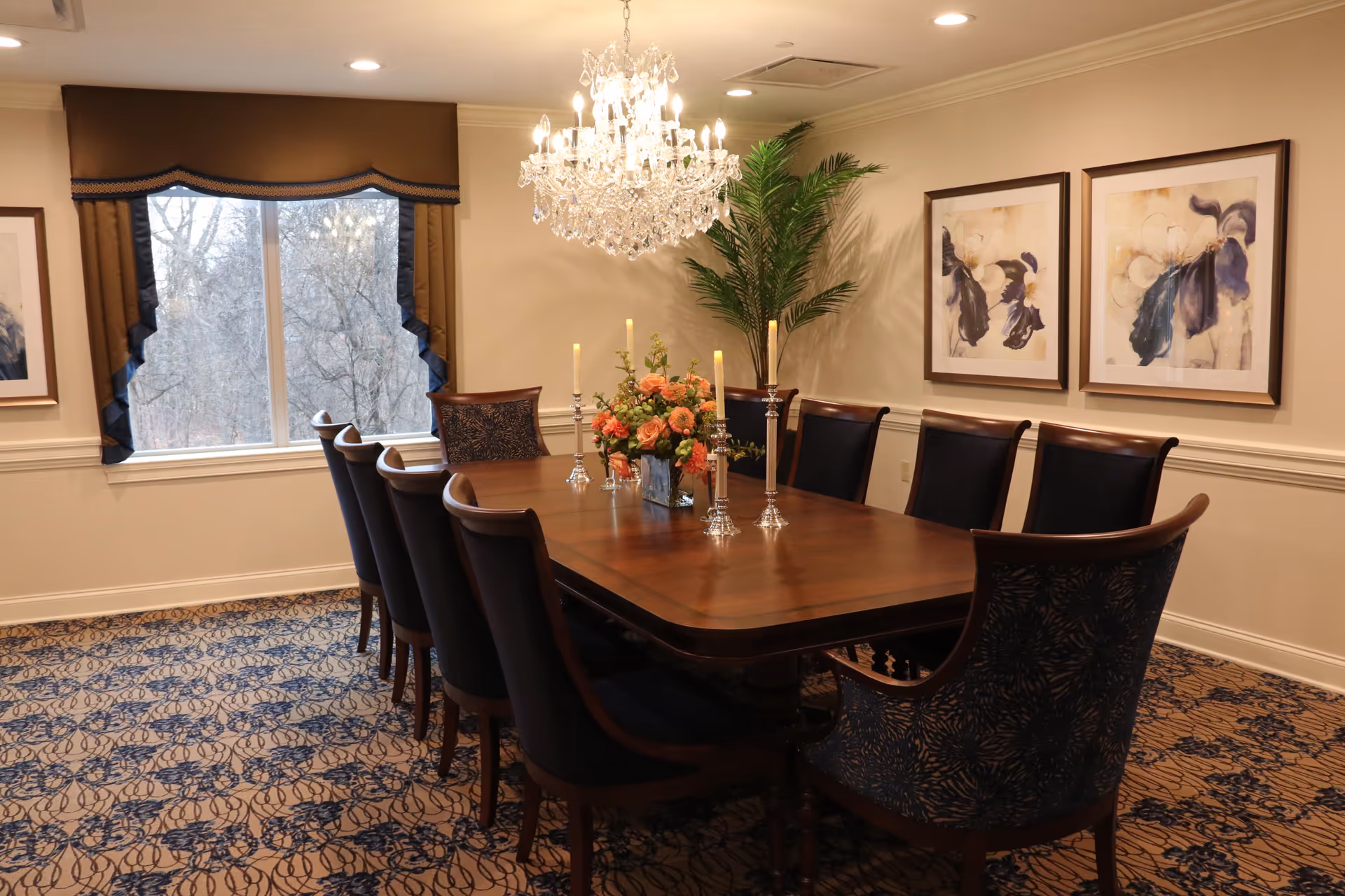 A formal dining room with a long wooden table surrounded by ten upholstered chairs. The table is decorated with a floral centerpiece and five tall candlesticks. A large crystal chandelier hangs above the table. The room has patterned carpet, beige walls with two framed floral paintings, a large window with brown and blue curtains, and a tall green plant in the corner.