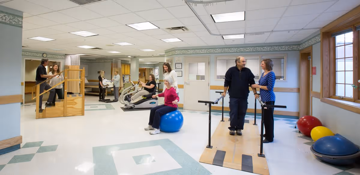 A rehabilitation or physical therapy room in a senior living facility with several elderly individuals engaged in various exercises. One woman is sitting on a blue exercise ball, a man is walking with support bars assisted by a therapist, and others are using exercise machines or walking up stairs with handrails. The room is well-lit with large windows and has a clean, spacious interior.