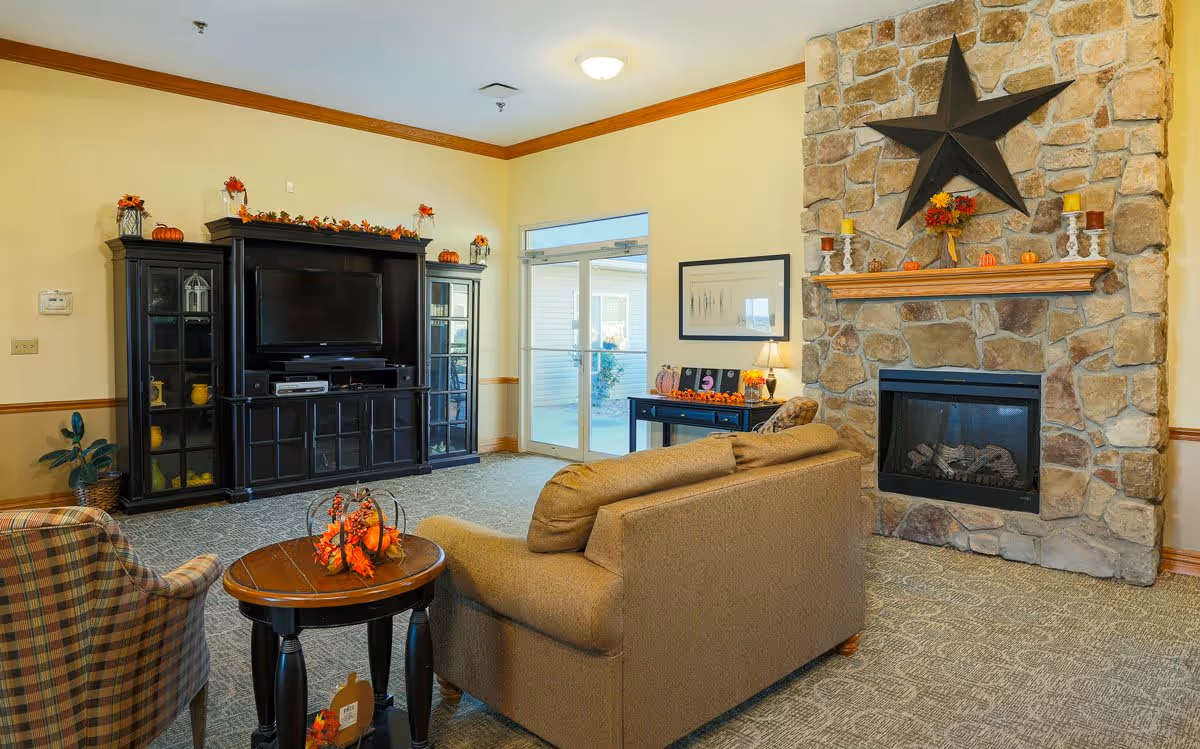 A cozy living room with a stone fireplace decorated with a large black star and autumn-themed items on the mantel. There is a brown couch facing a black entertainment center with a TV, flanked by glass cabinets. A small round wooden table with fall decorations and a plaid armchair are also visible. The room has beige walls with wooden trim and a carpeted floor.