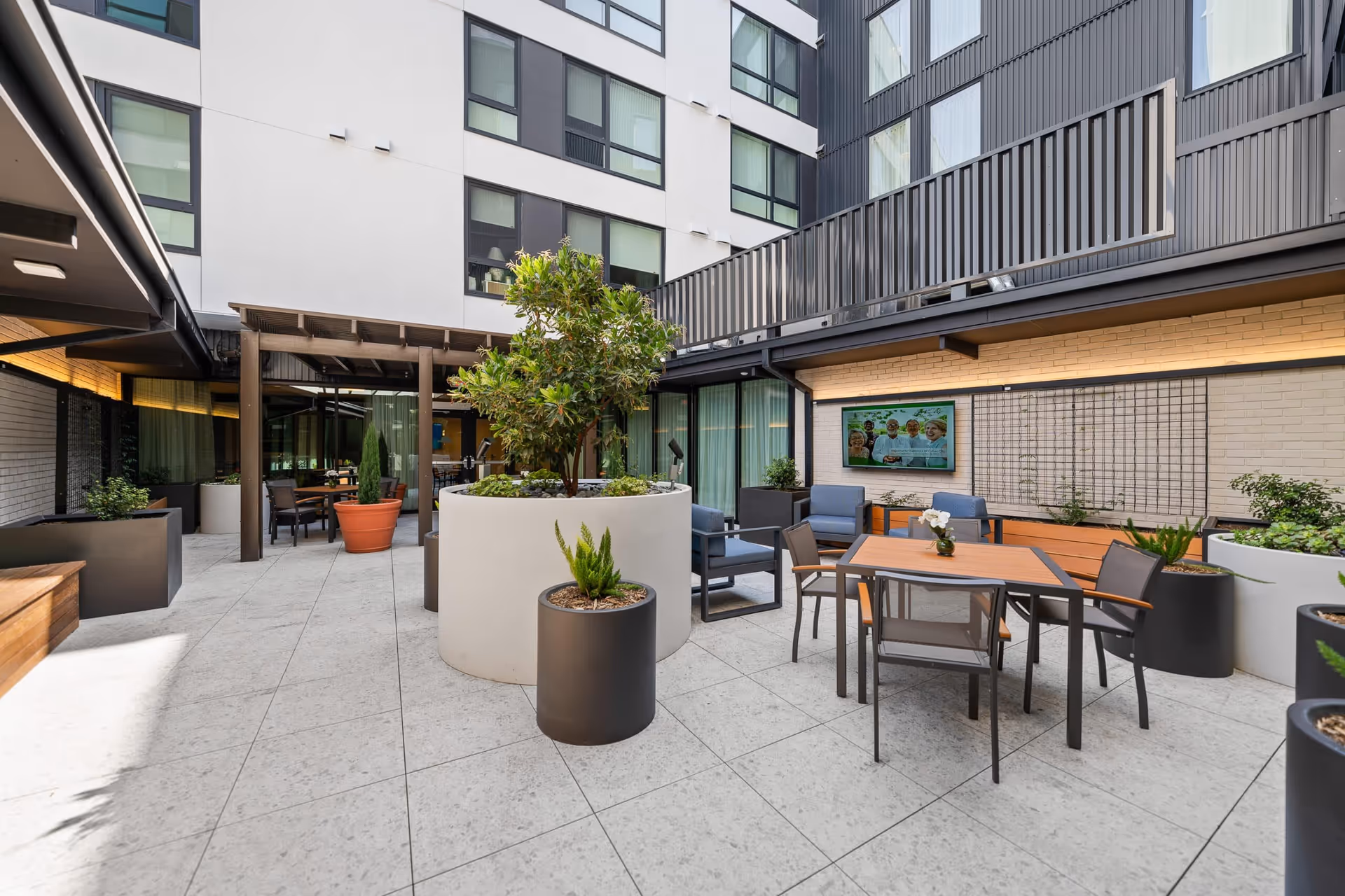 Outdoor courtyard area of a senior living facility with modern patio furniture including tables and chairs, large planters with greenery, a pergola, and a wall-mounted television displaying a group of smiling seniors. The space is surrounded by the building's windows and balconies.