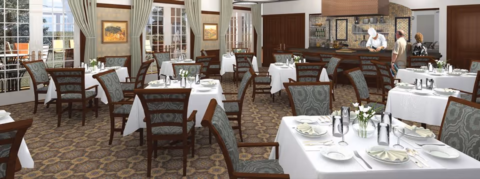 A spacious dining room with multiple tables covered in white tablecloths, each set with plates, napkins, silverware, and small flower arrangements. The room features patterned carpet, large windows with curtains, and a counter area where a chef is preparing food while two people stand nearby.