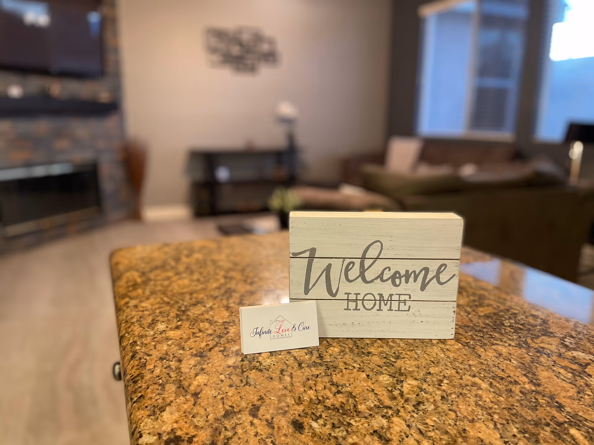 A close-up of a granite countertop with a small wooden sign that says 'Welcome Home' and a business card for Infinite Love & Care Homes. In the background, there is a blurred living room with a fireplace, a TV mounted on a stone wall, a couch, and a window.