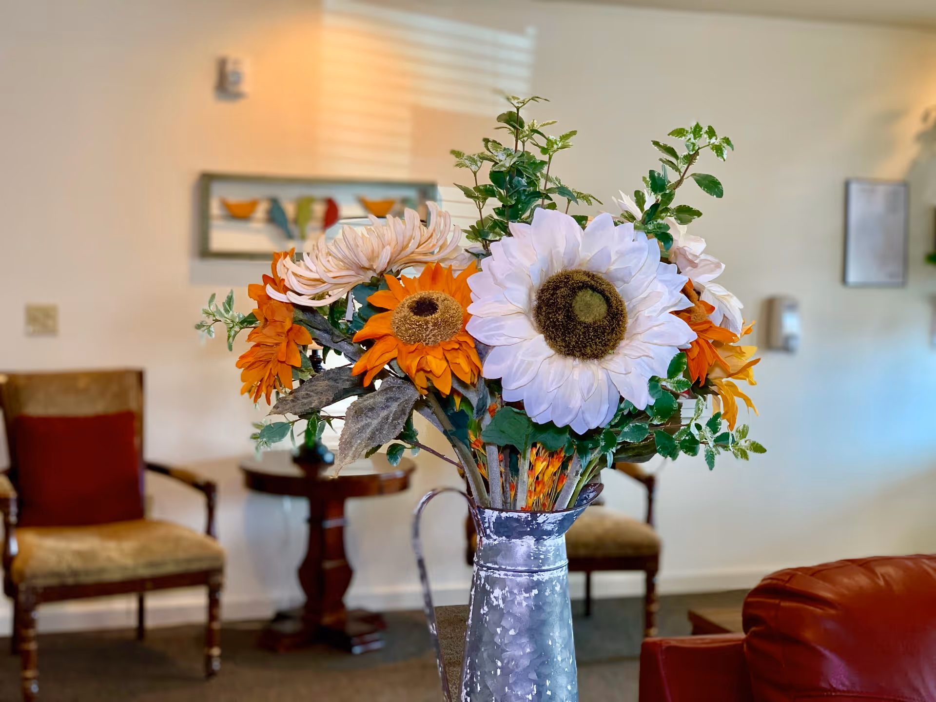 A close-up of a metal vase holding a bouquet of artificial flowers including large white and orange sunflowers, placed in a cozy living room with wooden chairs, a small round table, and wall decorations in the background.