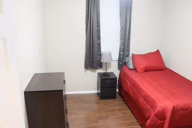 Small bedroom with a red twin bed, bedside table and lamp, dark dresser, and a window with gray curtains.