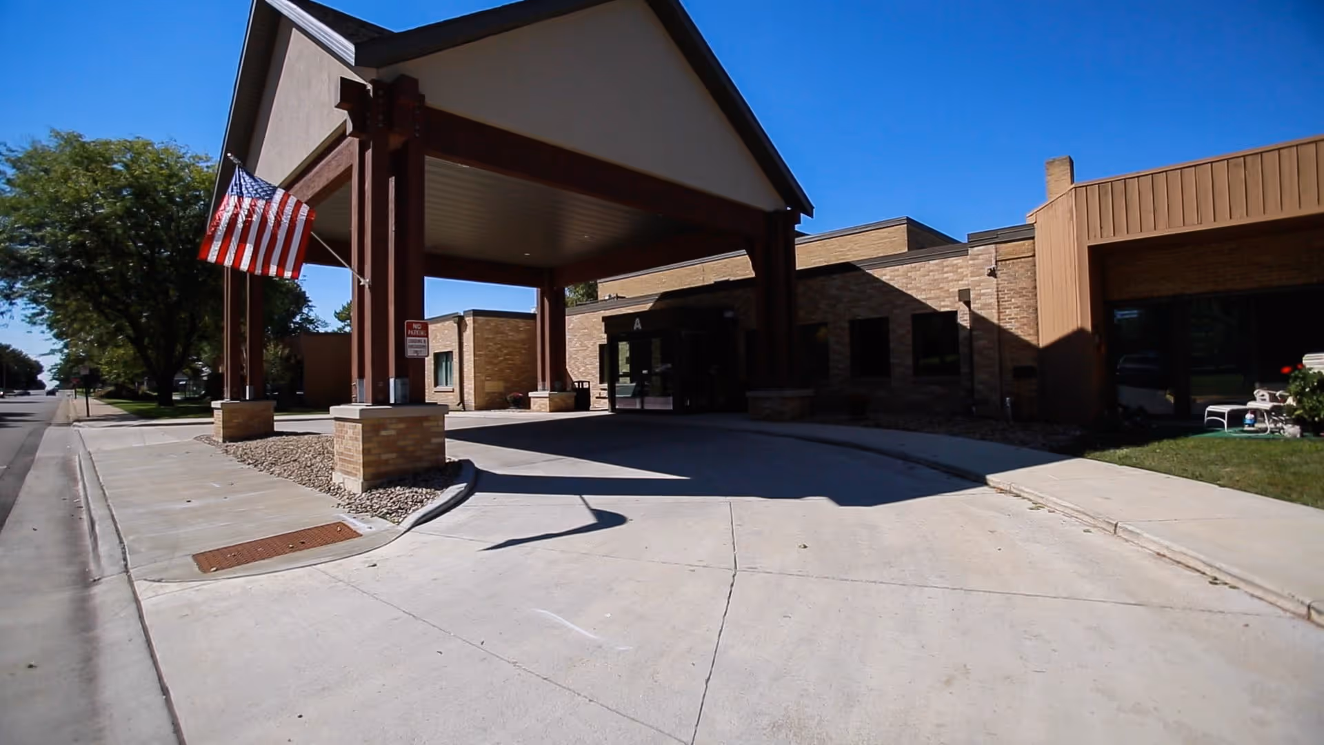 Entrance of Thorne Crest Senior Living Community with a covered driveway supported by wooden beams, an American flag hanging on the left side, and a brick building under a clear blue sky.