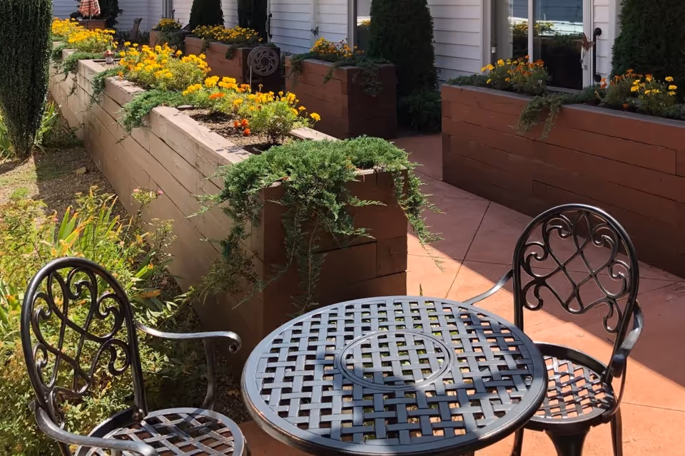 Outdoor patio area with a black metal round table and two matching chairs. Raised wooden flower beds with yellow and orange flowers and green plants line the walkway next to a white building with windows.