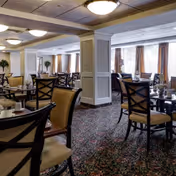 Dining room with multiple tables and chairs, patterned carpet, decorative columns, and large curtained windows.