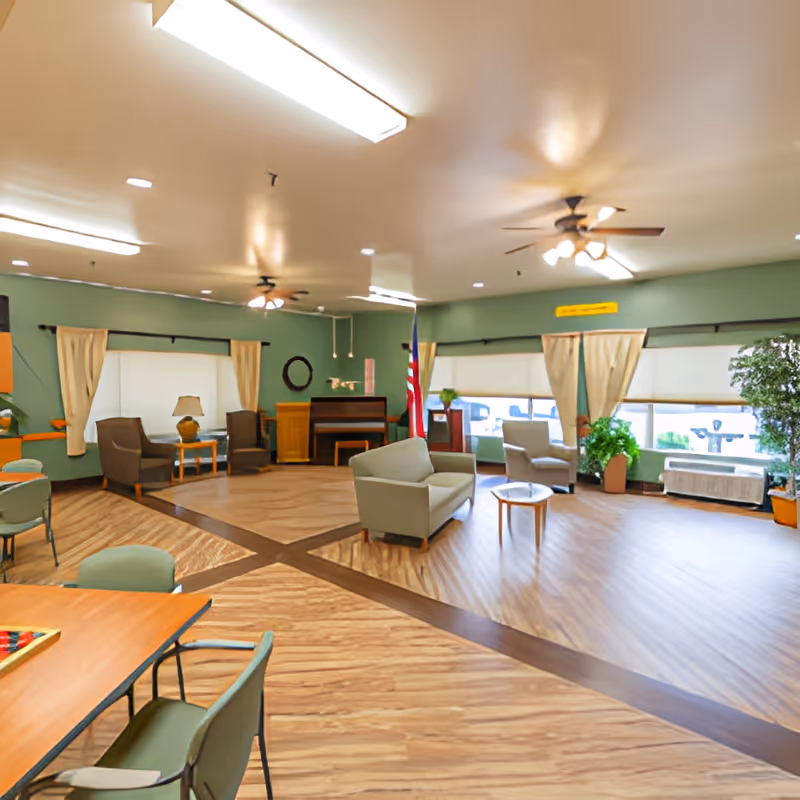 Spacious senior living common room with seating areas, tables, a piano, large windows and ceiling fans.