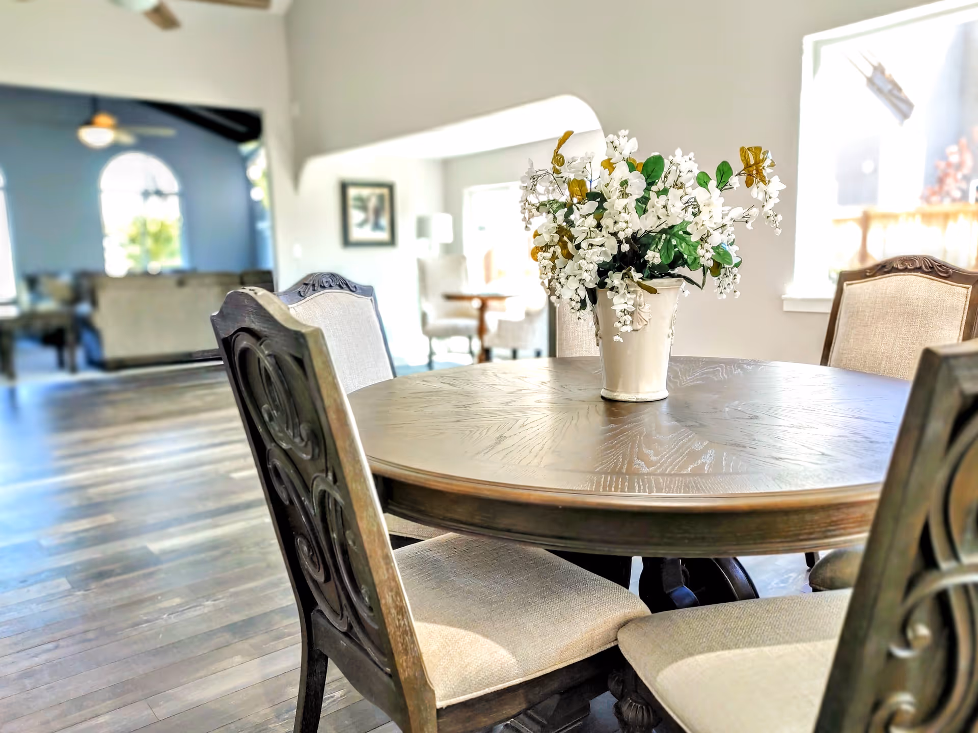 A round wooden dining table with a vase of white flowers in the center, surrounded by four upholstered chairs. The room has wooden flooring and an open layout with a living area visible in the background through an archway.
