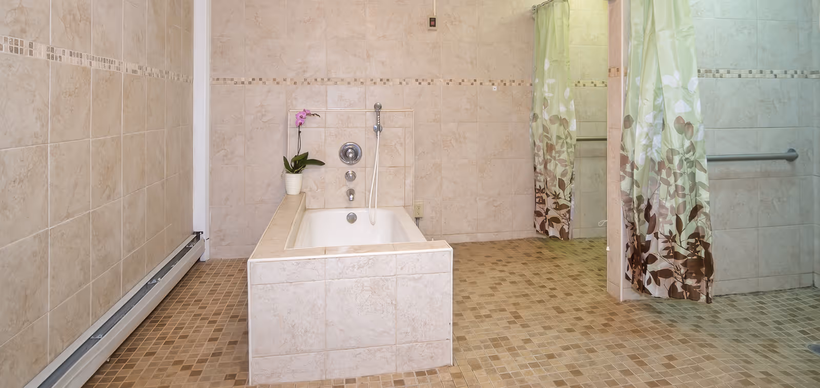 A spacious bathroom with beige tiled walls and floor. There is a built-in bathtub with a handheld showerhead and a potted orchid plant on the edge. To the right, there are two shower areas separated by a partial wall, each with a floral-patterned shower curtain and a grab bar on the wall.